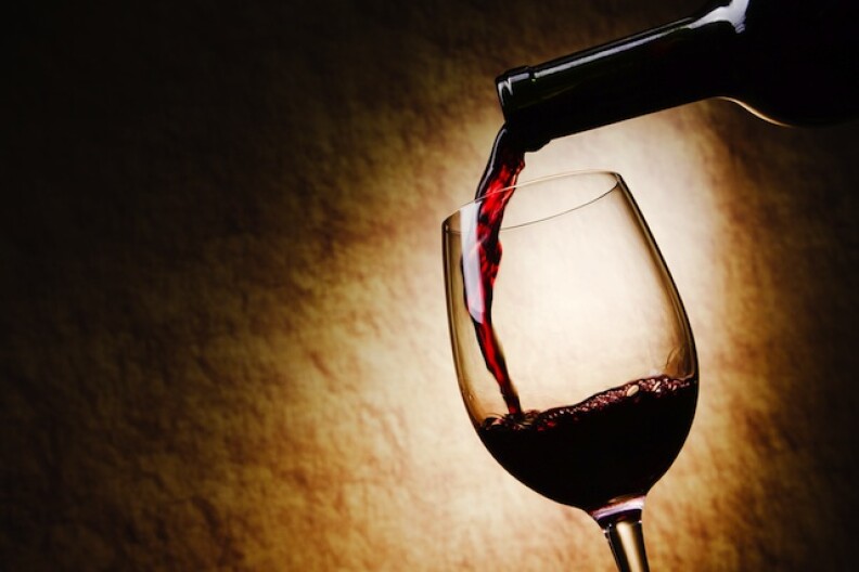 A bottle of red wine pours into a stemmed wine glass, shown in partial silhouette. The light in the image is more focused on the tan and brown gradient background.
