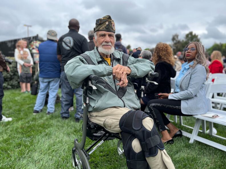 An older man with light skin tone sits in a chair and looks at the camera. He has braces on both of his knees. He's also wearing a hat that indicates he was in the military.