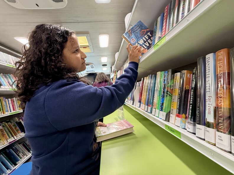 A girl wearing a navy blue sweater with long curly brown hair and medium light skin tone pulls a book from a white shelf.