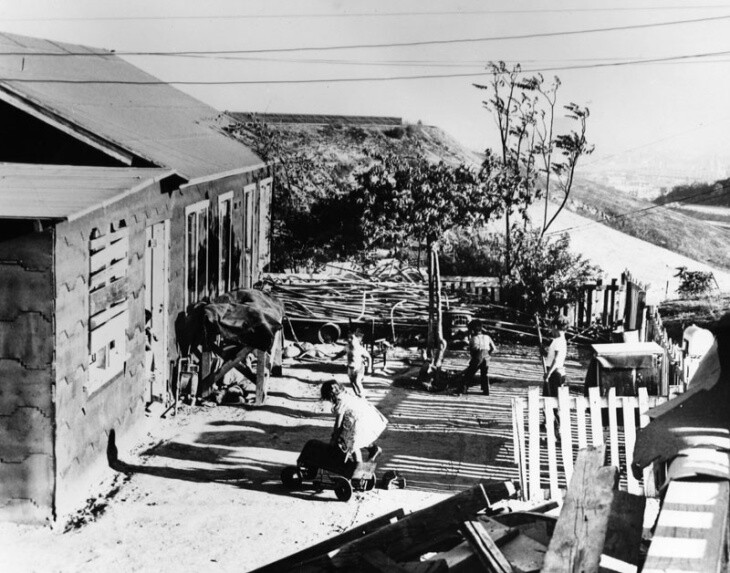 A black and white image of a ramshackle home on a hillside with people in the front lawn