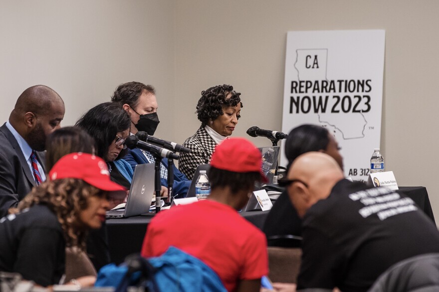 A group of people with light to dark skin tones are gathered in a room with table, mics and a sign that says: CA Reparations Now 2023