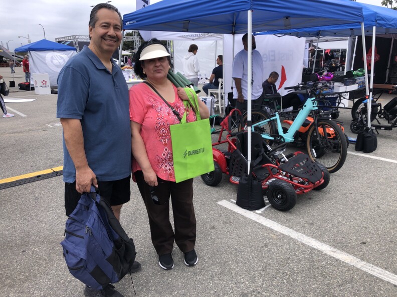 A man in a blue shirt holds a blue backpack beside and woman in a pink shirt holding a green tote bag. Electric go-karts and bikes are displayed behind them.