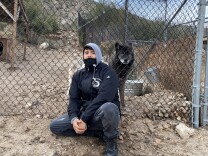 Wolf Connection team member Edward Amaya sits with hands clasped together. He wears a black jacket and grey hoodie. Beside him, behind a fence, sits his buddy Kenai, a black and brown male wolf who lives on the ranch. 