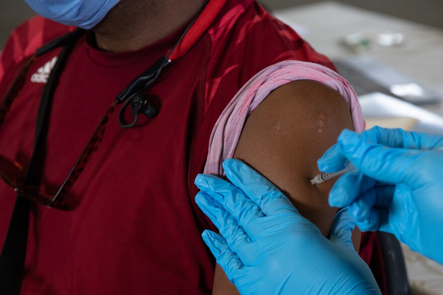 A person in blue medical gloves injects a needle into the upper arm of a person with dark skin, who is wearing a dark red athletic shirt.