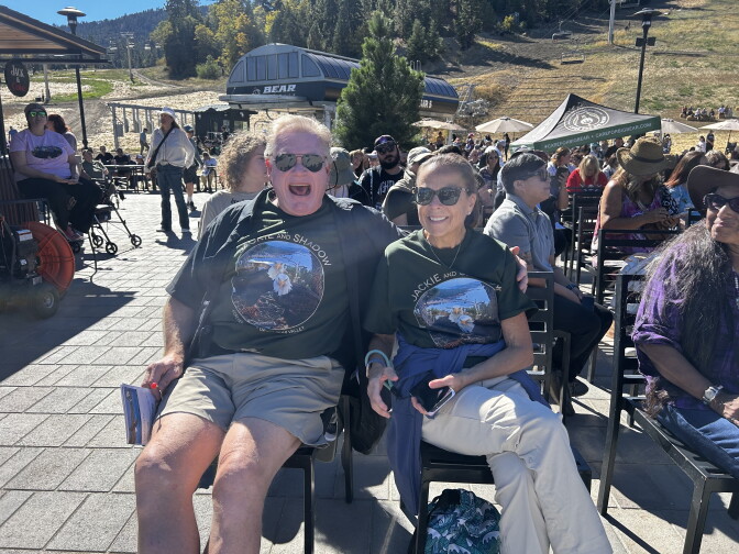 Two people wearing matching 'Jackie and Shadow' eagle shirts sit outdoors near a ski lift, smiling with a crowd behind them.