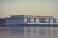 A large, low white building sits on the bank of a river. It is pictured from across the river.
