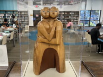 A wood sculpture of two people, they embrace and look at the viewer.