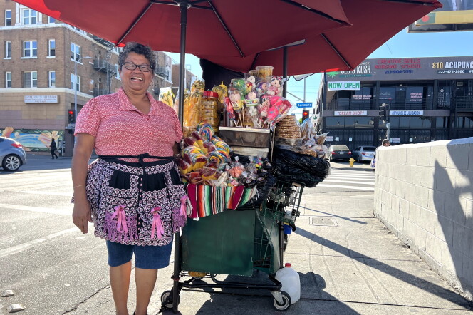 A Brown woman with short curly hair wearing a pink and black apron stand in front of a cart with an umbrella. On the cart, there are lollipops, an assortment of candies and chopped mangoes in plastic containers. 