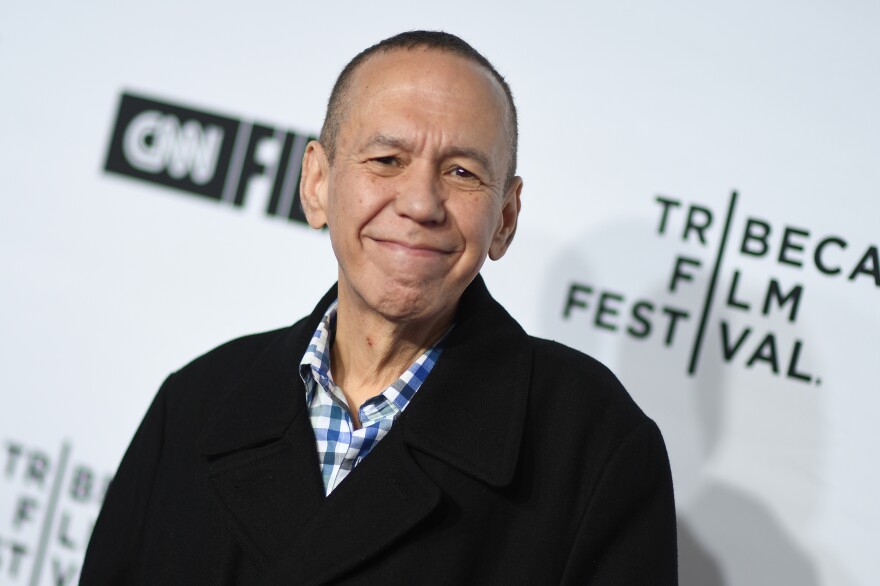 Gilbert Gottfried grins for the camera wearing a checked button-down shirt and black coat