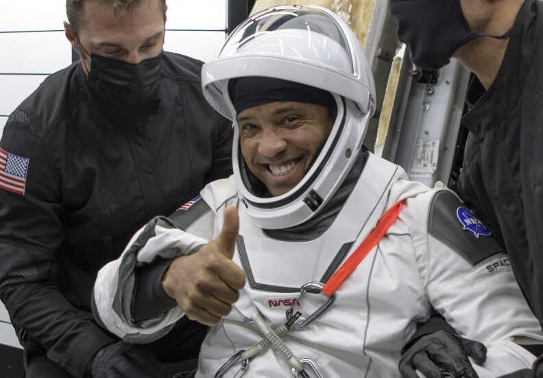 A Black man wearing a white spacesuit and helmet gives a thumbs up to the camera as two men in black coats wearing face masks assist him out of a large metal spacecraft