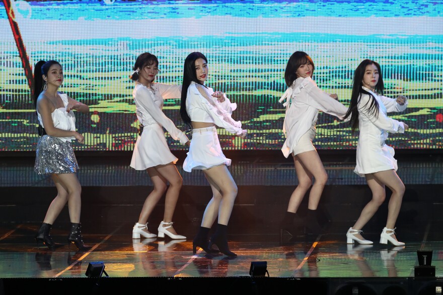 A K-pop band onstage, dancing. Five young Asian women who appear to be in their late teens or early 20s are all turned to face the right of the frame, with their knees slightly bent and their arms in motion. They wear all white, with short skirts and long sleeved tops. Their faces are turned towards the camera.