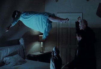 A young girl, Linda Blair, floats above a bed in a small bedroom. Blair wears a white night gown. Her arms are outstretched , her legs slightly together as she looks like she's laying down but in the air. Two men in all black stand at the foot of the bed, Max von Sydow and Jason Miller. von Sydow has one arm raised above his head with something in it. The room is dark, the only light coming from a lamp on the nightstand next to the bed. The room has a bluish glow to everything. 