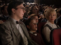 A light-skinned man with glasses. 1950's jacket and bowtie sits in a packed movie theater next to a young light-skinned boy wearing a sweater and a bowtie. Next to him is a light-skinned woman with a blond bob and bangs. She's looking at the man. The young boy is looking entranced at the screen.