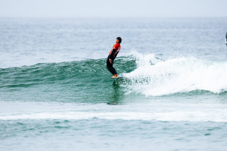 A man in a red rash guard and wetsuit leans back on the nose of a longboard while riding a small wave to shore.