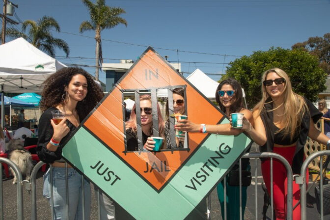 Five women hold drinks outdoors while standing near an oversized Monopoly jail square. 