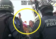A yellow circle is focused on a man with light-toned skin and hair holding what appears to be a phone high above his head. In the foreground two officers in U.S. Capitol Police gear have their backs to a body-worn camera capturing the scene