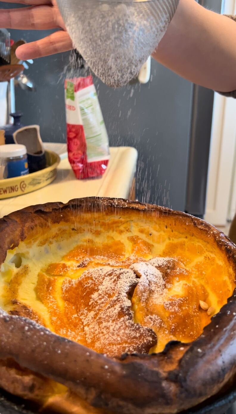 A pair of fair-skinned hands holds a mesh strainer while adding powder to an oversized pancake.