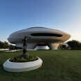 A saucer-shaped building sits on a lawn behind a bronze statue of a figure holding a pole atop a pedestal.