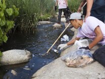A volunteer in a pink shirt and gray hat reaches over to pick out a bottle from the LA River.