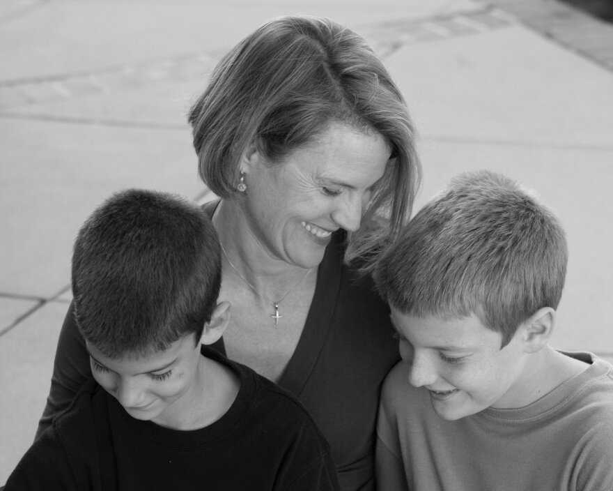 A woman with light tone skin and hair smiles as she has her arms around two boys. She wears a cross necklace.