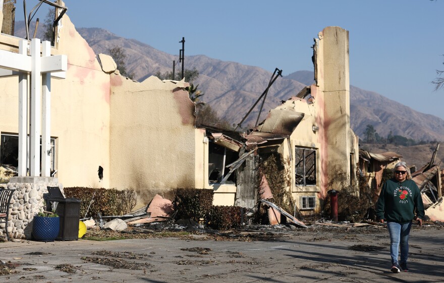A partially burned down building with beige walls. Mountains can be seen in the background and a person wearing a green sweatshirt and jeans walks by.