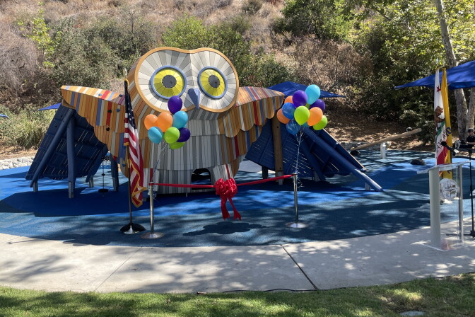 A 12-foot owl-shaped playground is seen on a rubber mat surface, surrounded by grass, balloons and vegetation.