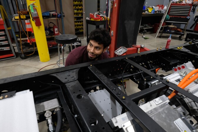 A young man with light brown skin and a maroon long-slevved shirt and dark brown wavy short hair smiles as he lifts equipment onto a chassi of a truck. 