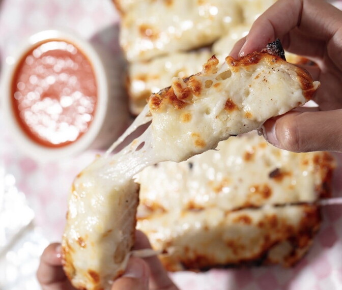 A hand pulls apart a piece of cheesy, golden-brown garlic bread over a basket lined with checkered paper, with a small bowl of marinara sauce in the background.