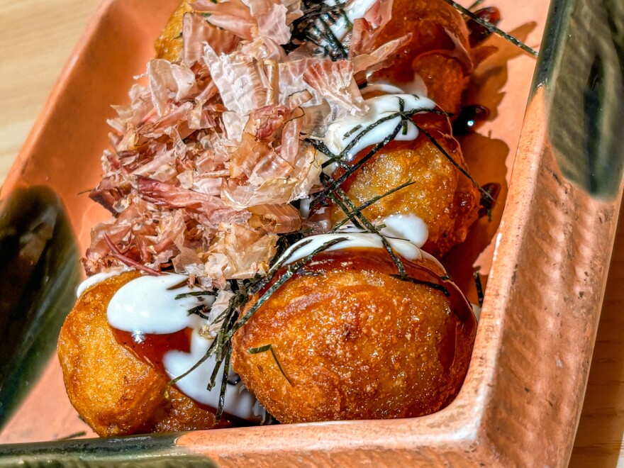 A close up of round fried balls covered in white and brown sauces topped with thin strips of seaweed in a pinkish-colored dish.