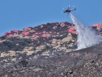 A helicopter sprays fire retardant on areas that show signs of burning. A red substance can be seen on the hills in the background.