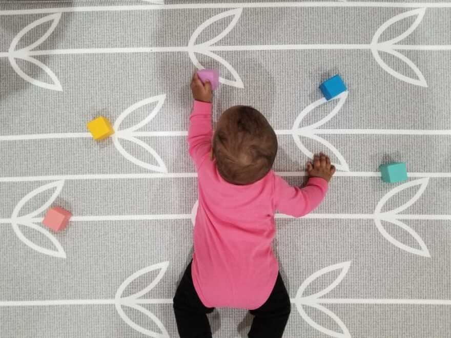An image shot from above of a medium skin-toned baby in a pink long-sleeved onesie on her belly on a gray carpet. She is surrounded by five colored blocks and reaches out to the purple one in front of her.
