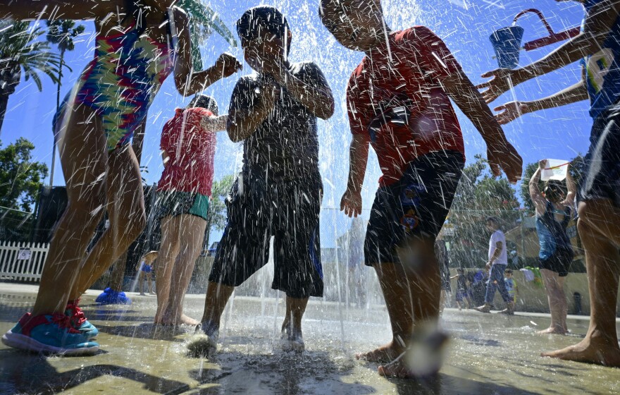 A group of children in shadows get soaked by a water at a splash pad.