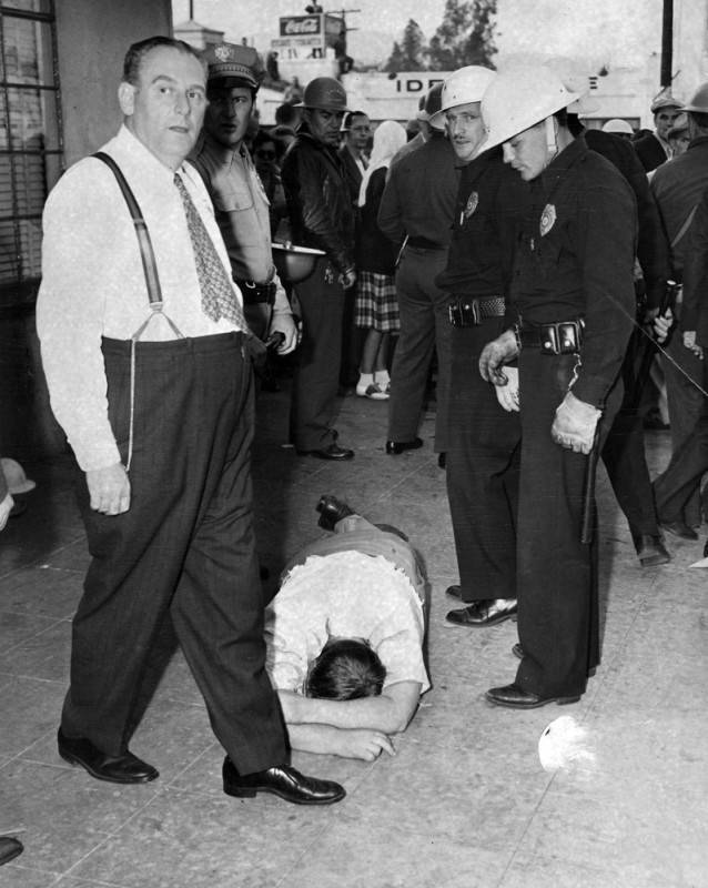 Two policemen in hard hats are standing over the man and several other people can be seen in the background.