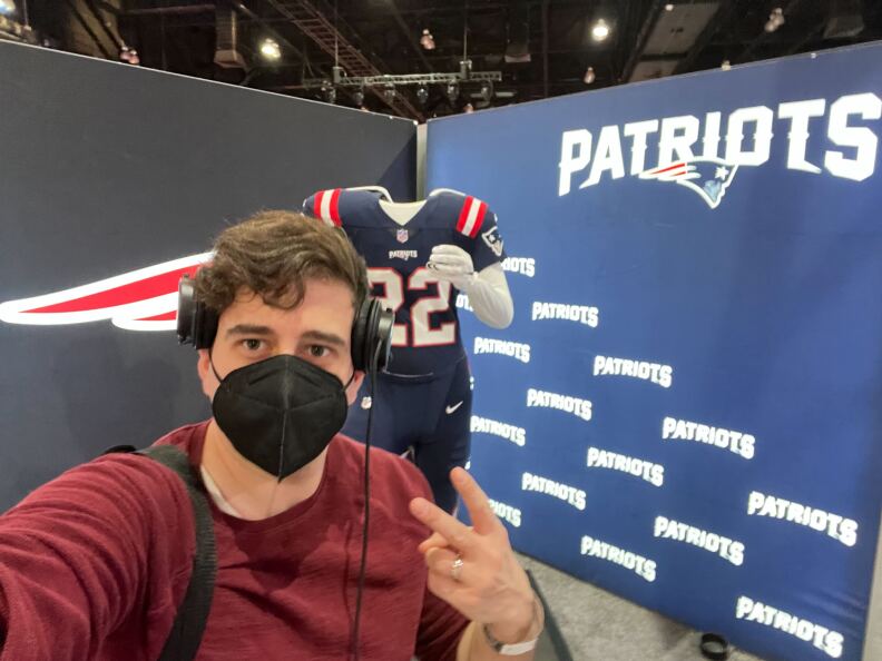 Reporter Matt Dangelantonio, wearing a black face mask and and radio headphones, poses in front of of a New England Patriots jersey at the NFL Super Bowl Experience.