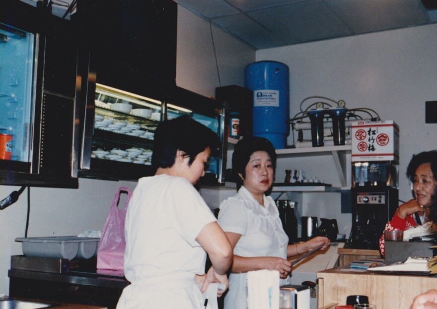 A photo of two Asian women — one is looking at the camera while the other has her back to the lens — in white working at a restaurant counter, talking to another Asian woman