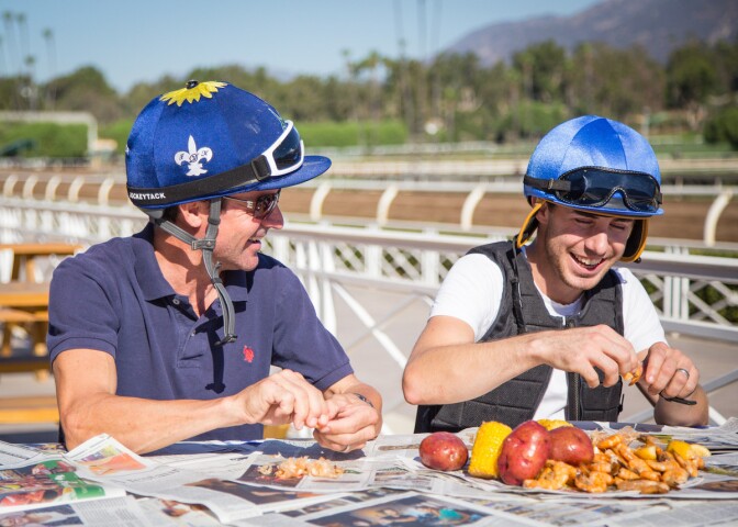 Two jockeys eat a shrimp boil trackside at Santa Anita Park. The shrimp, corn and potatoes are atop of newspapers.