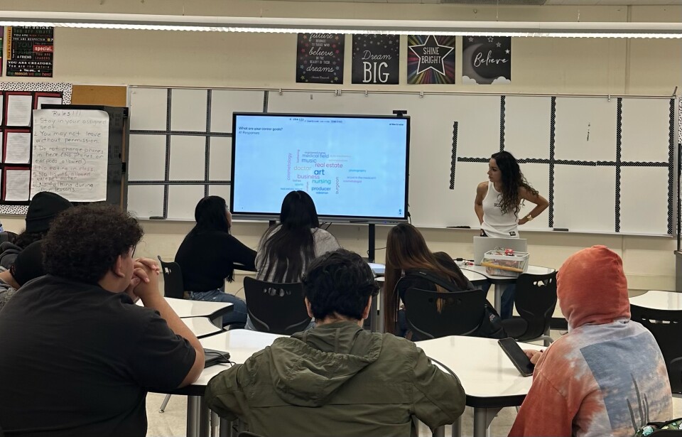Person with long dark hair in front of a classroom with people at desks looking at her