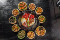 A large grey-colored stone dish containing a variety of meats and vegetables sits in the center of an assortment of round, light brown-colored steamer baskets, each containing different types of food on a black surface. 
