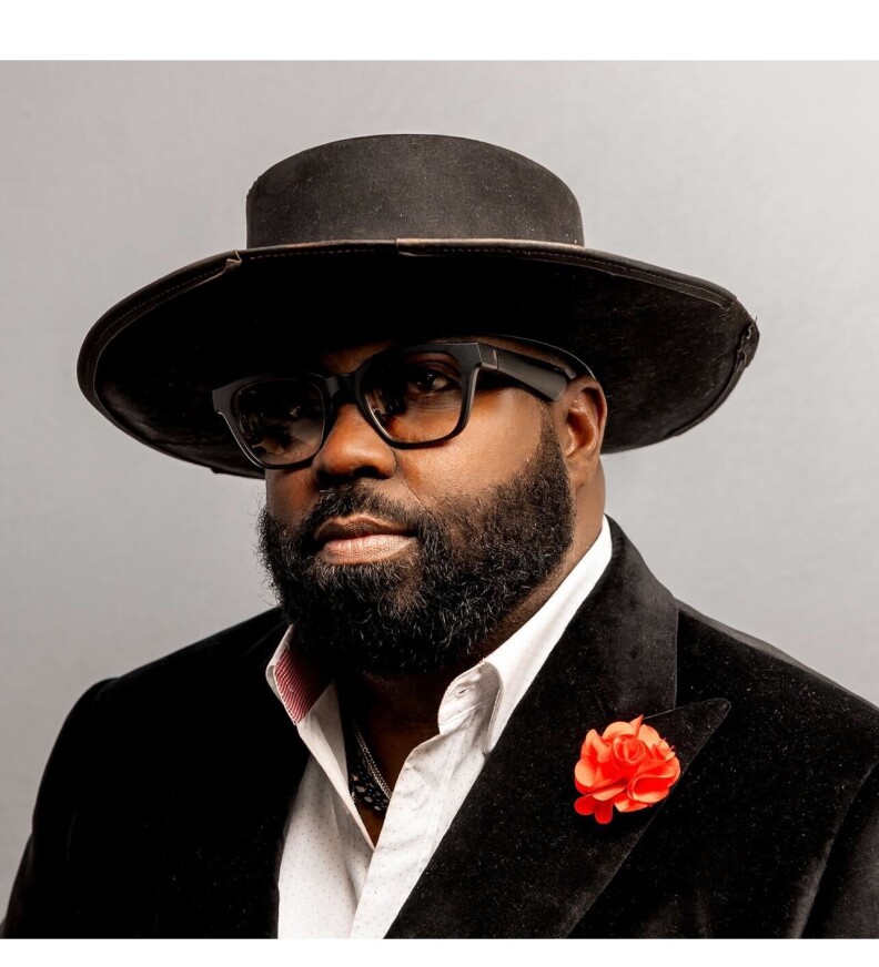 A black man with a short beard wearing a wide-brimmed black hat, black-rimmed a glasses and a velvet black suit with a red flower pinned to the left lapel in a portrait. 