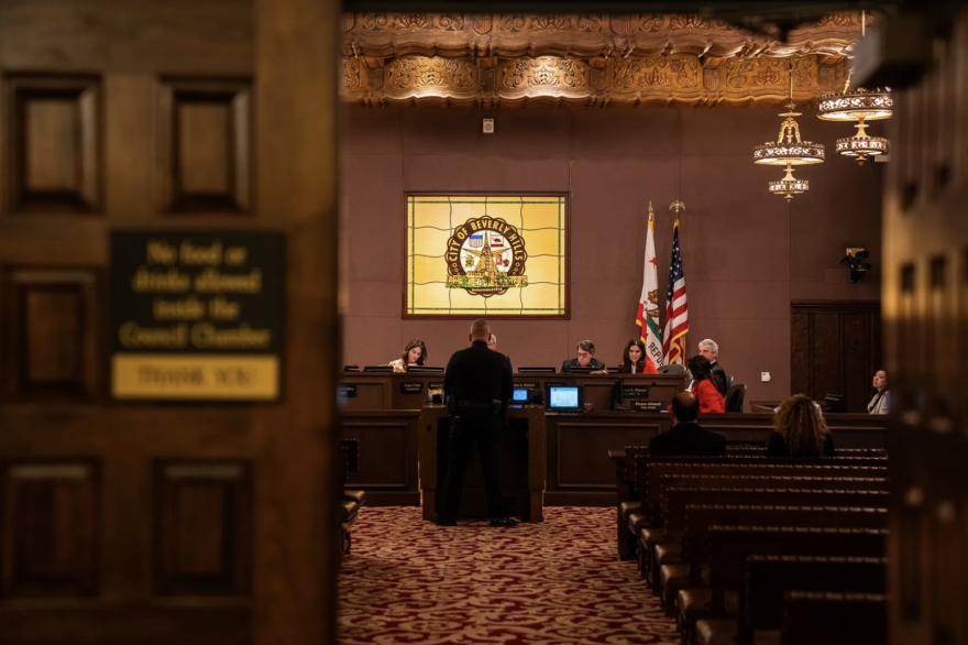 People inside the city council chambers of Beverly Hills can be seen from just outside of the doorway. Some sit in the rows of seats in the room, while one person stands before a panel of others, speaking at the podium, back turned to the camera.