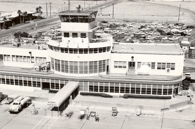 A black and white photo shows a white building flanked with bay windows and a tower on top. Cars are seen in the distance in a parking lot with the tale of an airplane seen in the lower left corner. 