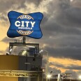A big blue sign that say "City Brewing & Beverage Irwindale" is seen at sunset. Lights illuminate the sign. Large clouds can be seen behind it. Palm trees and power lines are seen in the right hand corner.
