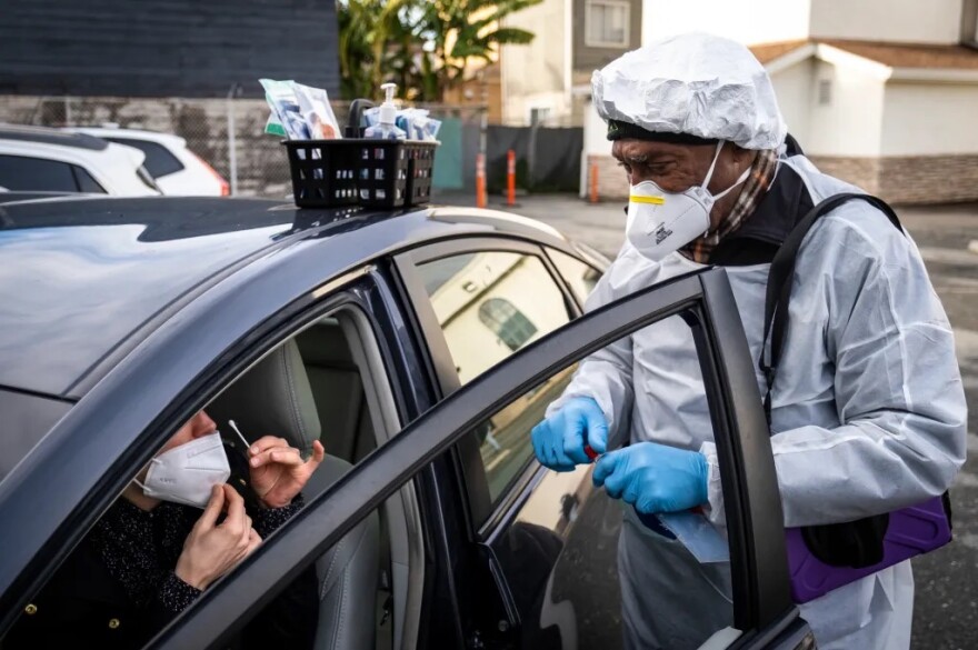 A person in a light blue smock, wearing blue gloves, a white mask, and a head covering, with a portion of a COVID test in his hand and a purple bag over his shoulder, stands inside the open door of a car, the window down. In the car, a woman whose face is partially obscured wears a white mask below her nose and holds a white swab. There is a black basket on top of the black car that contains some sort of soap/hand sanitizer dispenser, along with a number of other items. A white and brown building is in the background, as well as what appears to be a fence, some palm trees, orange stanchions, other vehicles, and a larger white and off-brown building.