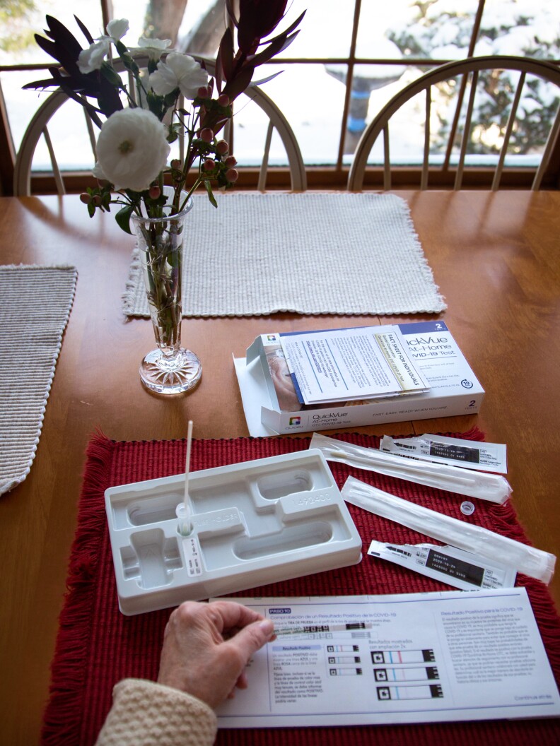 A table in a home has an open test kit spread out. A person holds a test,
