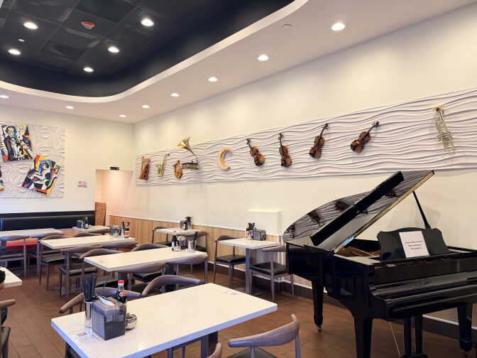 White tables and wooden chairs fill a dining room, next to a black baby grand piano with its lid propped open. A wavy white wall above displays violins, a saxophone, a trumpet, a harp and a gramophone.