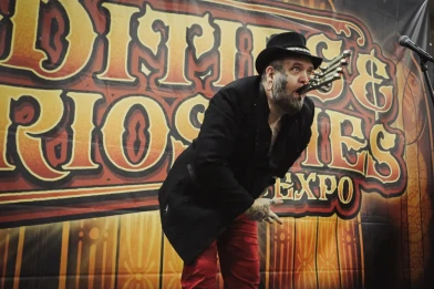 A light-skinned man with a beard and black hat holds a strange metal contraption in his mouth. 