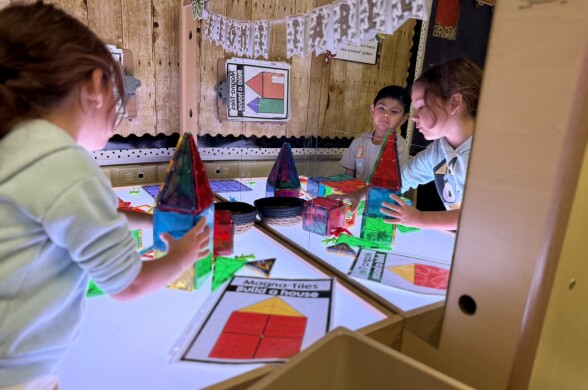 Two students with medium light skin tone construct shapes out of clear, blue, red, yellow and green plastic tiles on a lit surface