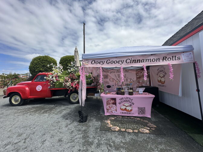 Photo of a truck next to a pop-up business. The pop-up business had a banner titled "Ooey Gooey Cinnamon Rolls." Another banner, on top of a table, has a label saying "The Little Sugarplum."