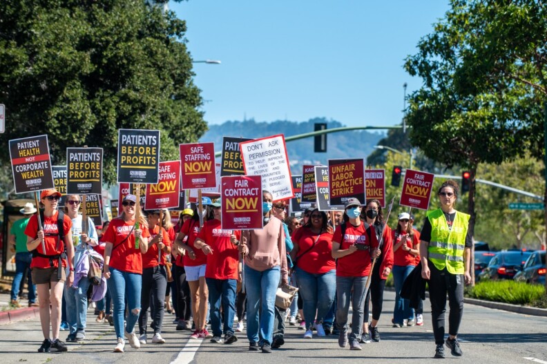 More than a dozen Kaiser mental health workers march down a street in Oakland as part of a more than three week strike. They wear mostly read t-shirts. Their signs read "Patients Before Profits" and "Fair Contract Now"