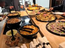 A man with facial hair wears a dark suit while bowing and gesturing over multiple plates of food at a table.